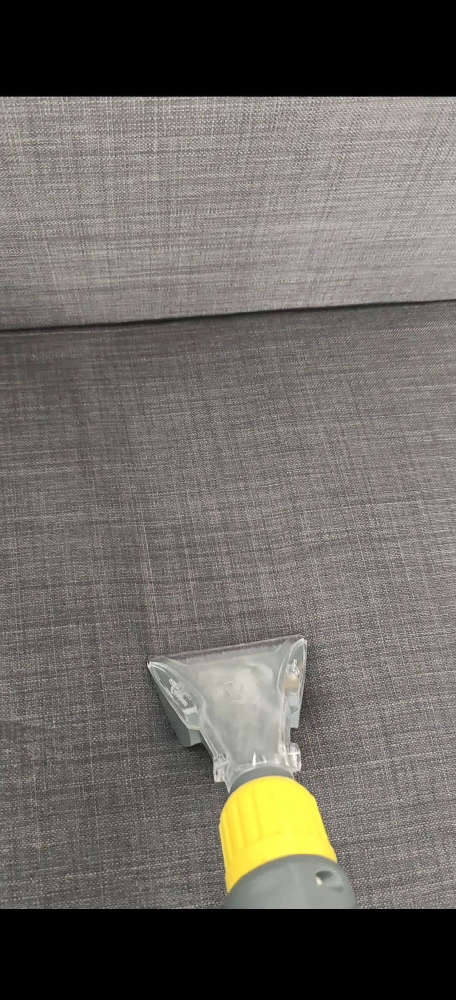 Dysza odkurzacza piorącego na szarej, tekstylnej kanapie. Widoczne detale struktury materiału i żółty element łączący dyszę z rurą.