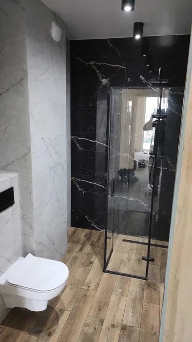 Wykończona łazienka z kabiną prysznicową typu walk-in, płytkami imitującymi marmur na ścianach i drewnopodobną podłogą. Widok na minimalistyczną toaletę i czarne oświetlenie sufitowe.