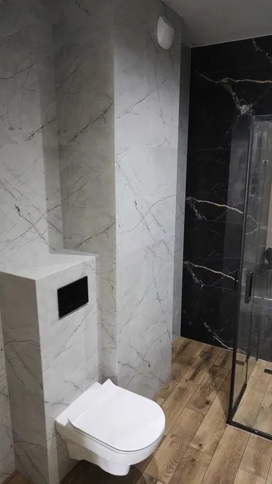 Nowoczesna łazienka z marmurowymi płytkami, podwieszaną toaletą i drewnopodobną podłogą. Kabina prysznicowa z czarnymi ramami.