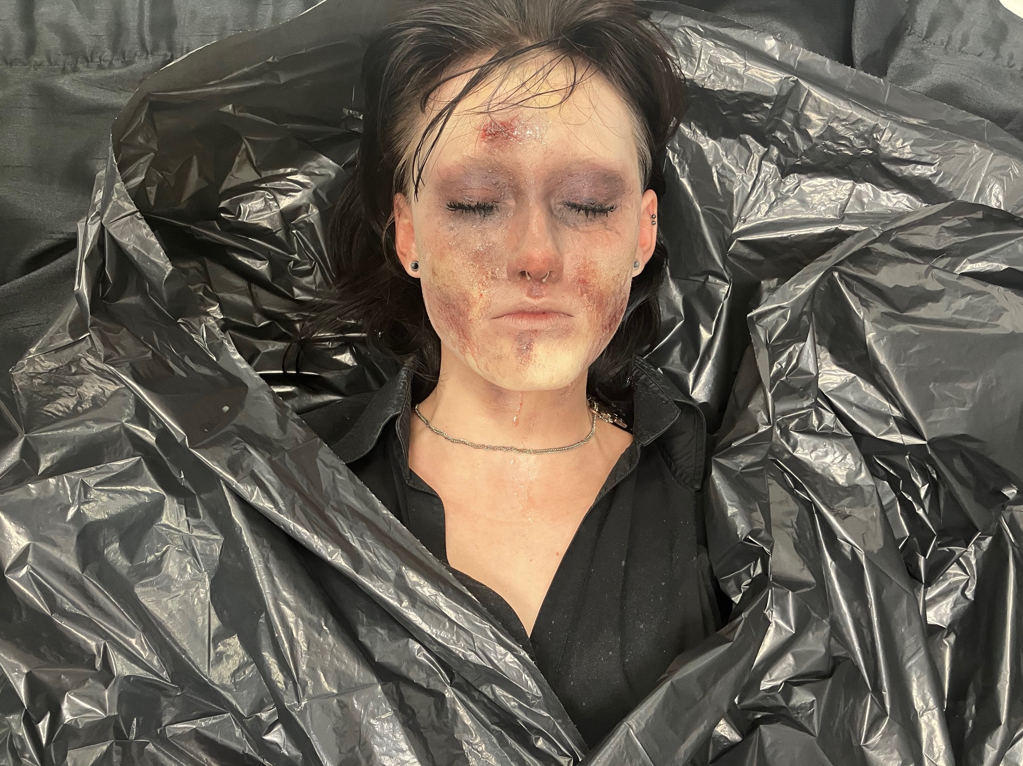Efekt makijażu charakteryzatorskiego we Wrocławiu: kobieta z zamkniętymi oczami, realistyczne rany i siniaki na twarzy, otoczona czarną folią. Intensywne, artystyczne wykonanie.