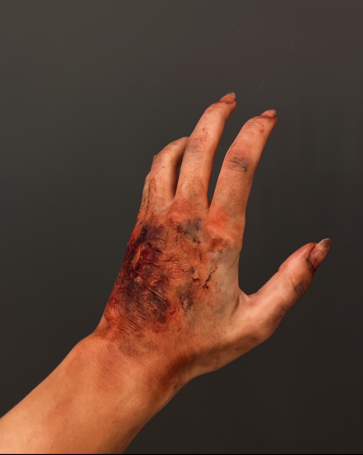 Dłoń z realistycznym makijażem efektów specjalnych: rany, siniaki i zabrudzenia, na tle szarego studia, ukazująca umiejętności w charakteryzacji filmowej.