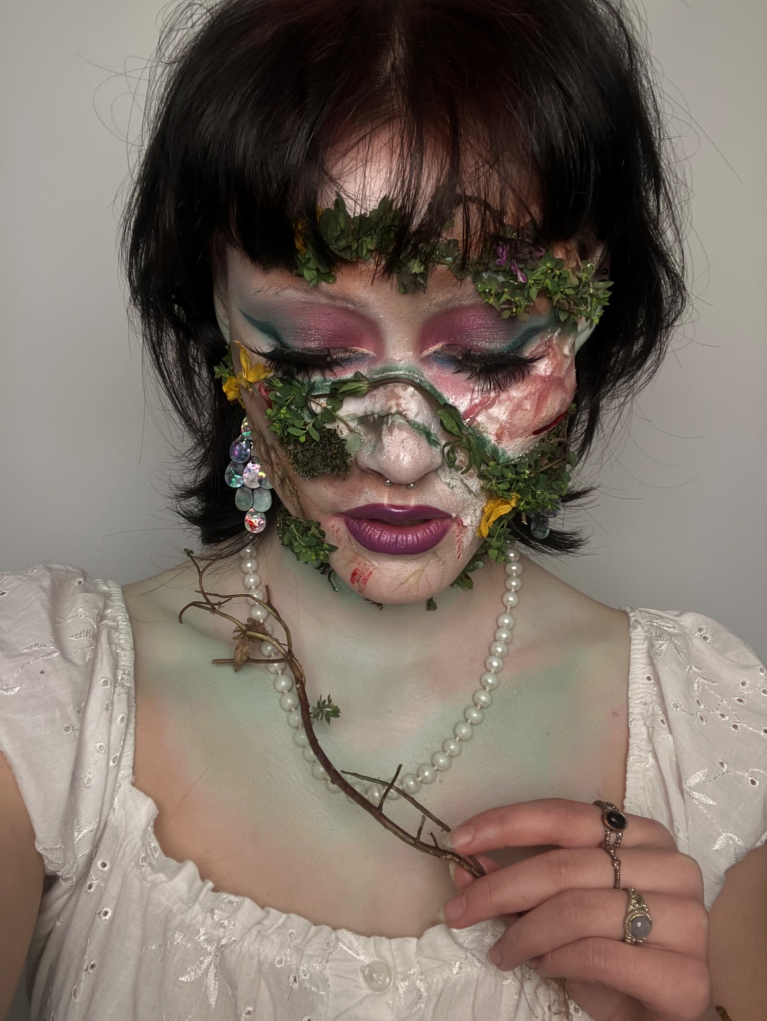Kreatywny makijaż artystyczny z elementami roślinnymi na twarzy kobiety, z zamkniętymi oczami i delikatnym oświetleniem, akcentujący unikalny styl i precyzję wykonania.