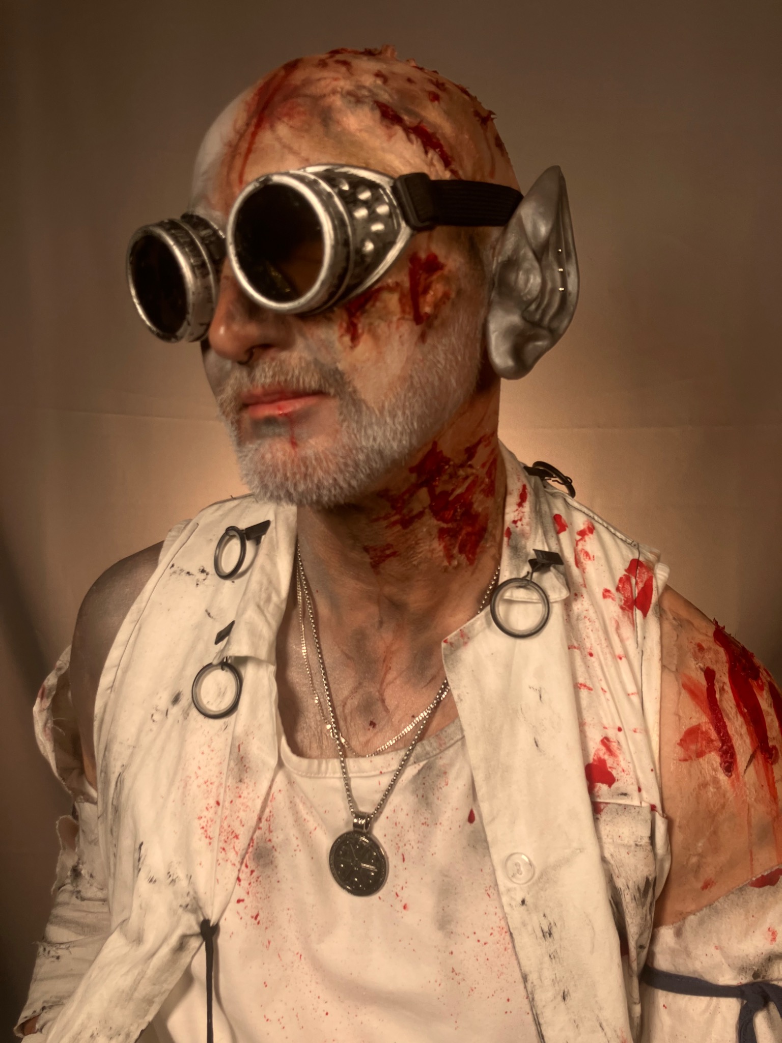 Mężczyzna z charakteryzacją: sztuczne rany, gogle steampunk, elfi uszy. Realistyczny makijaż na Halloween lub cosplay, na beżowym tle.