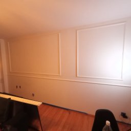 Tomasz Murach - Białe ściany z dekoracyjnymi listwami w kształcie prostokątów, zabezpieczone niebieską taśmą malarską przy podłodze. W tle widoczne oświetlenie i elementy wyposażenia.