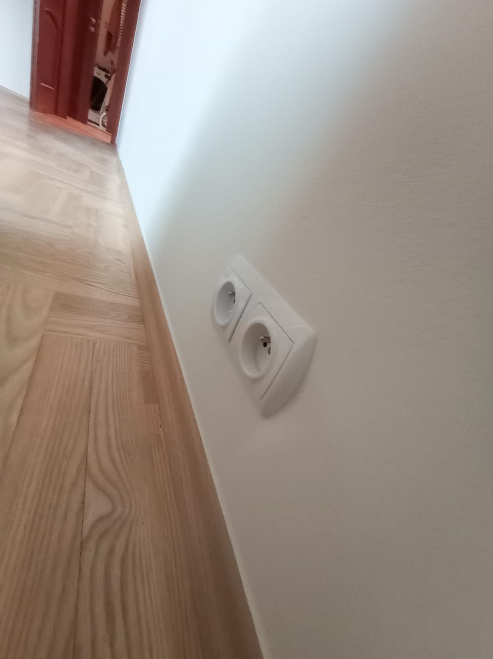 Białe, podwójne gniazdko elektryczne na świeżo pomalowanej, gładkiej ścianie w jasnym odcieniu, widoczne fragmenty parkietu i drzwi w tle. Ujęcie z dołu.