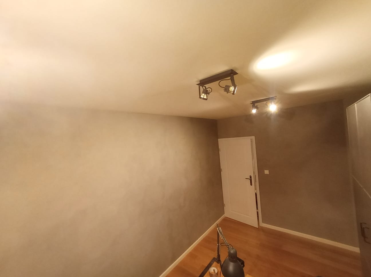 Pokój po szpachlowaniu i malowaniu na szaro, z drewnianą podłogą, białymi drzwiami i lampami sufitowymi. Widoczna lampka biurkowa w rogu.
