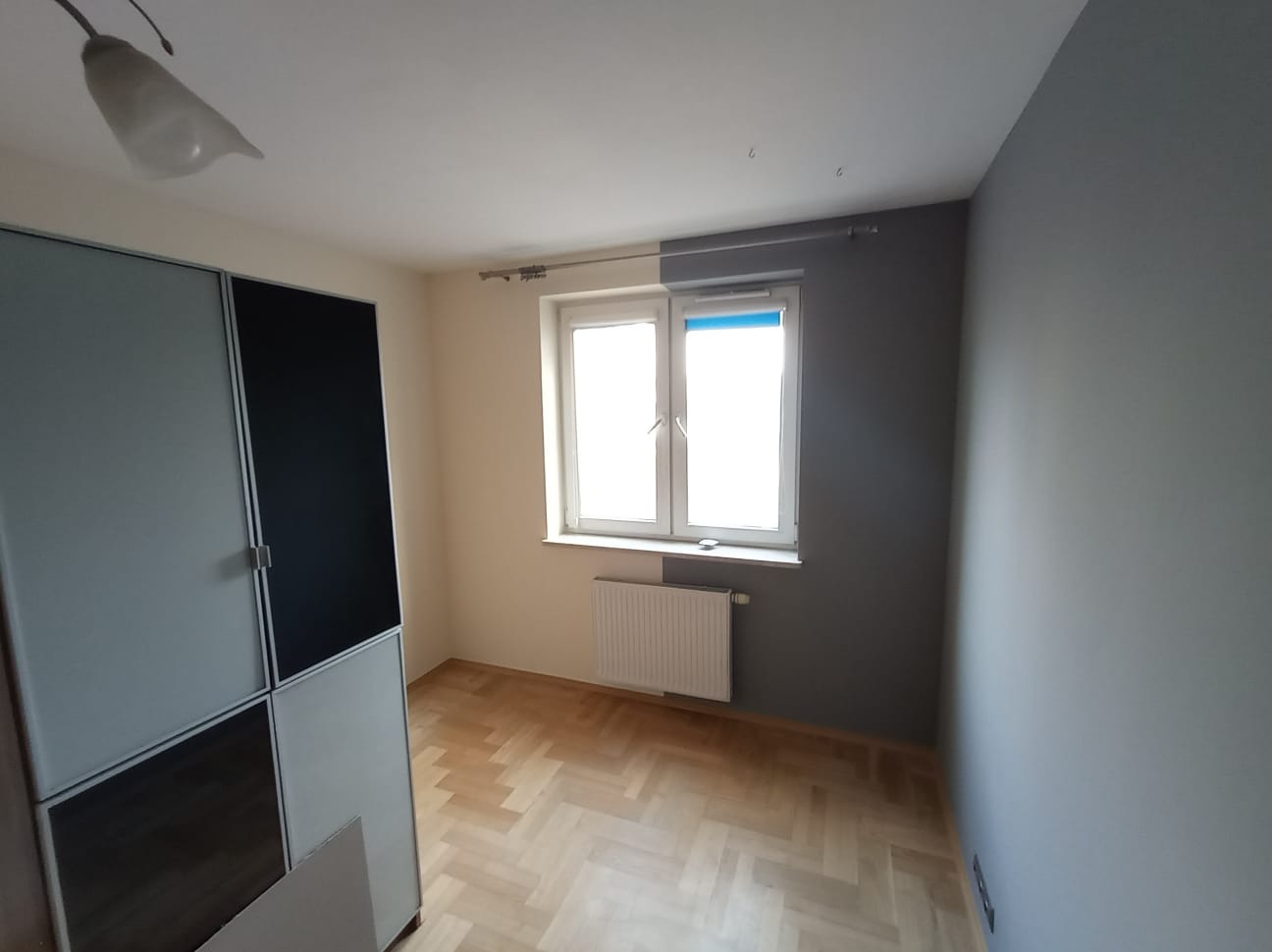 Pokój po remoncie: ściany pomalowane na szaro i kremowo, parkiet w jodełkę, okno z niebieską roletą, nowoczesna szafa z czarnymi elementami. Widok na odnowione wnętrze.