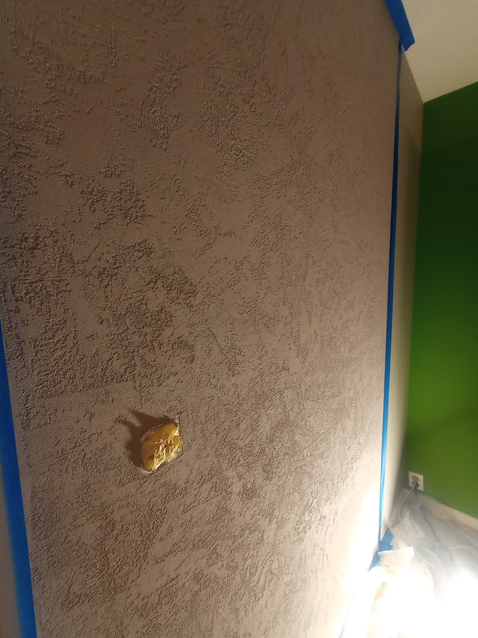 Ściana z nałożoną strukturą, zabezpieczona taśmą malarską i folią. Widoczne gniazdko elektryczne zabezpieczone folią. Ściana pomalowana na szaro-beżowy kolor.