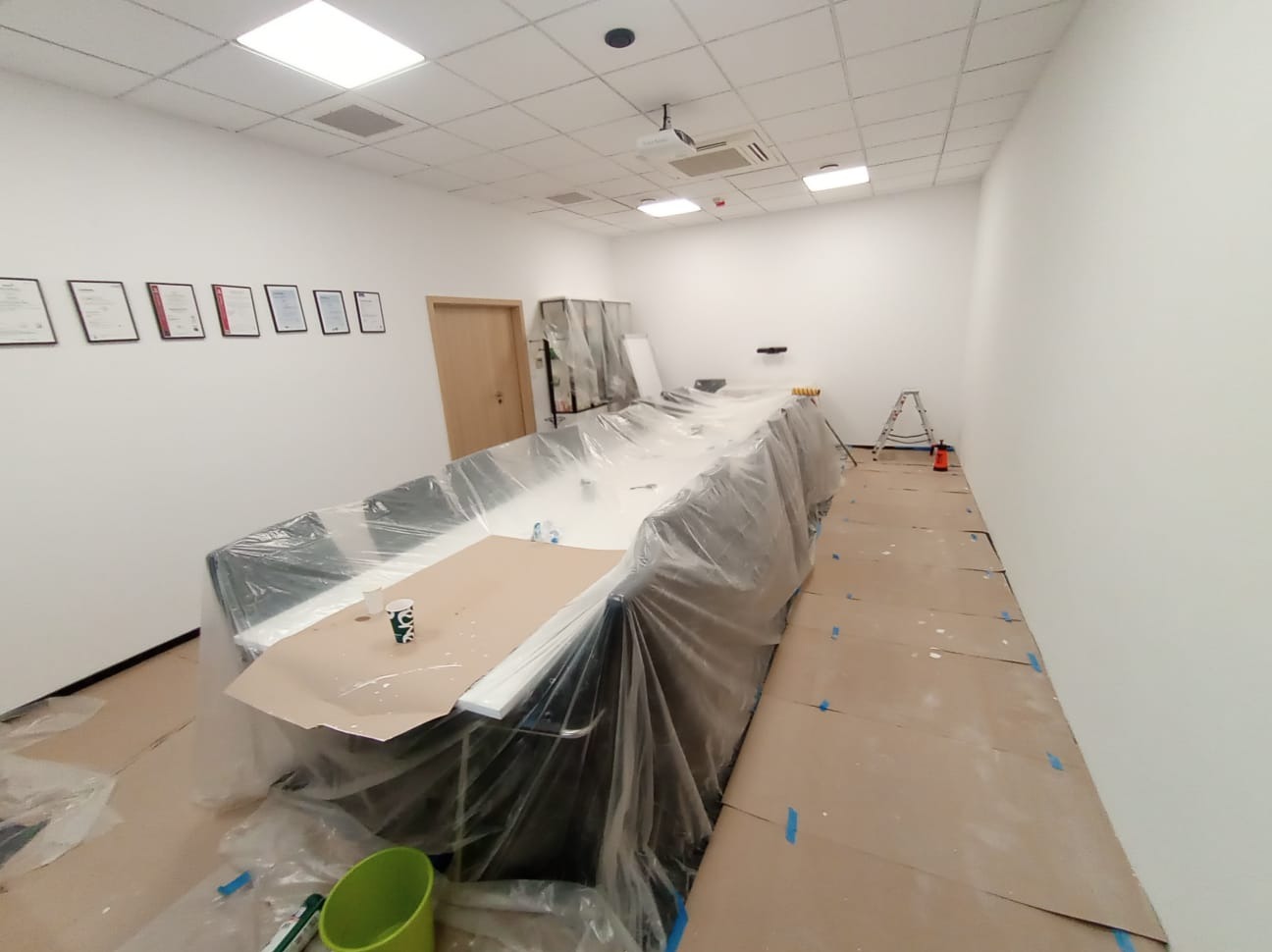 Białe biuro przed malowaniem: meble zabezpieczone folią, podłoga kartonem. Widoczna drabina i akcesoria malarskie. Przygotowanie do malowania biura.