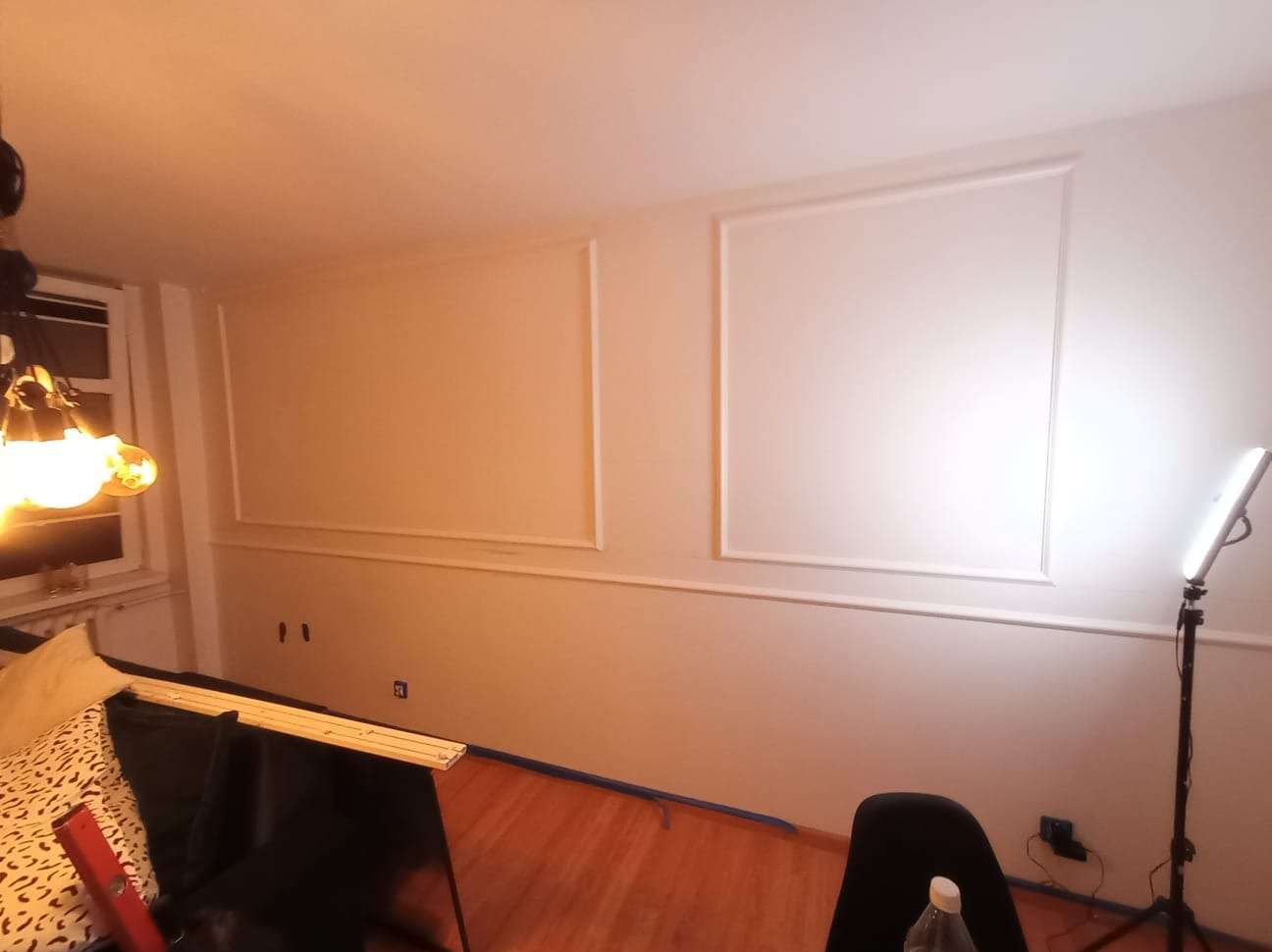 Białe ściany z dekoracyjnymi listwami w kształcie prostokątów, zabezpieczone niebieską taśmą malarską przy podłodze. W tle widoczne oświetlenie i elementy wyposażenia.