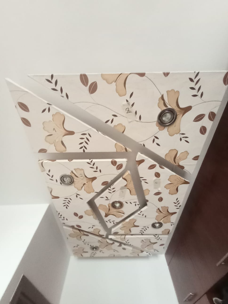 Dekoracyjny sufit podwieszany z oświetleniem punktowym i wzorem kwiatowym w odcieniach beżu i brązu, widziany pod kątem, z fragmentem ściany i szafki w tle.