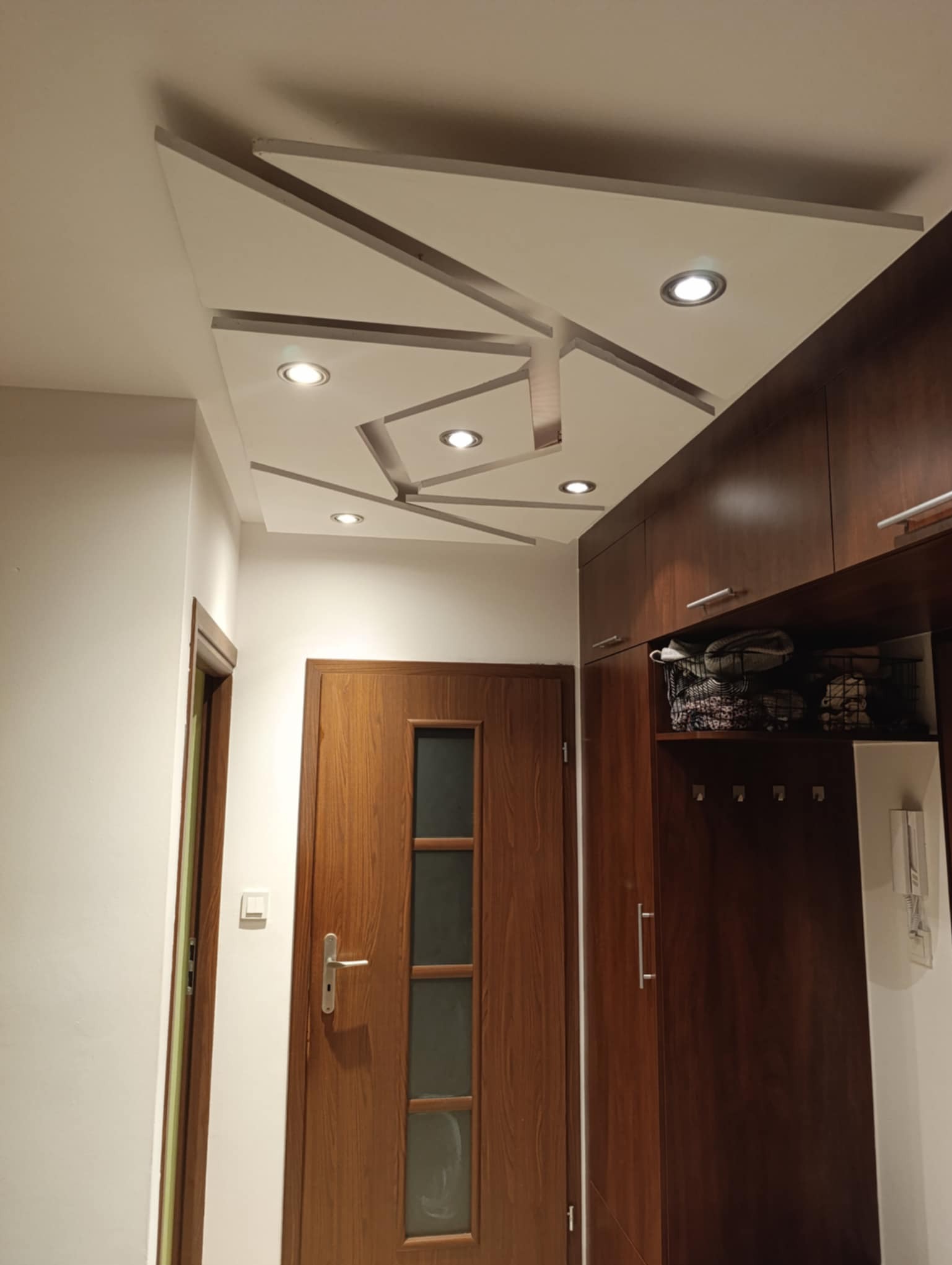 Nowoczesny przedpokój z drewnianymi drzwiami i szafą w zabudowie. Sufit podwieszany z geometrycznym wzorem i oświetleniem punktowym. Białe ściany, widoczny fragment włącznika.
