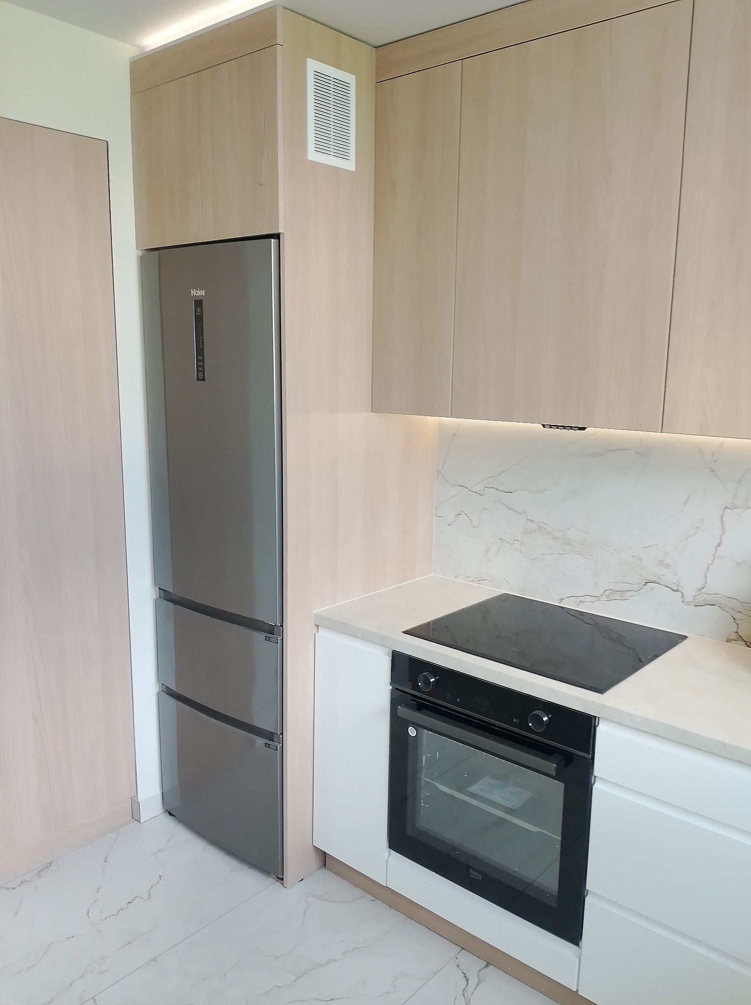 Jasna kuchnia z zabudową meblową: lodówka Haier, płyta indukcyjna, piekarnik Beko. Białe fronty, marmurowy backsplash i podłoga. Nowoczesny design.