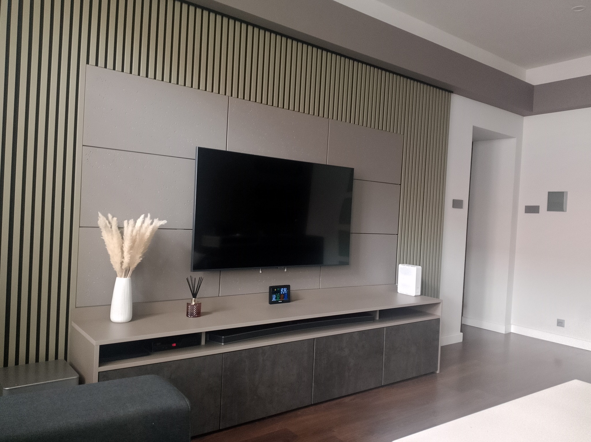 Nowoczesna aranżacja ściany TV z lamelami i szarą zabudową meblową. Minimalistyczny design, dekoracje w postaci trawy pampasowej i dyfuzora zapachowego.