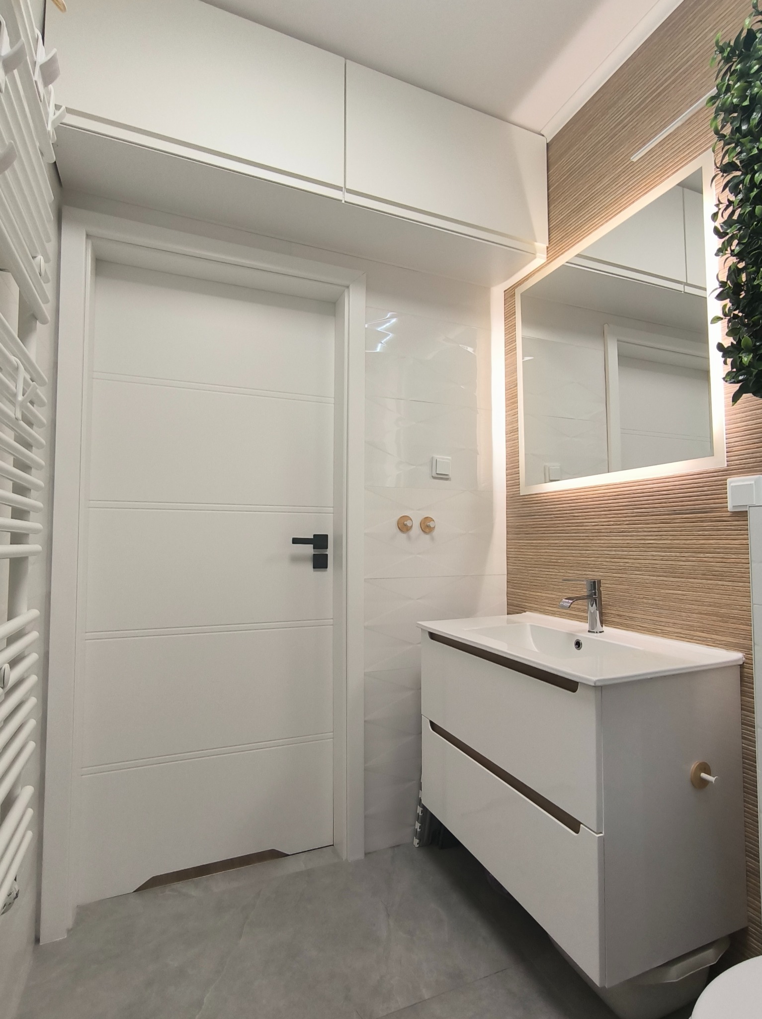 Nowoczesna łazienka po remoncie: biała szafka z umywalką, podświetlane lustro, białe płytki i drzwi z czarną klamką. Minimalistyczny design z drewnianymi akcentami.