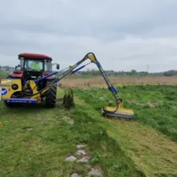 Czerwony traktor z wysięgnikiem i kosiarką rotacyjną Marolin M580SX na zielonym polu po koszeniu. Widoczne ślady koszenia i fragmenty trawy.