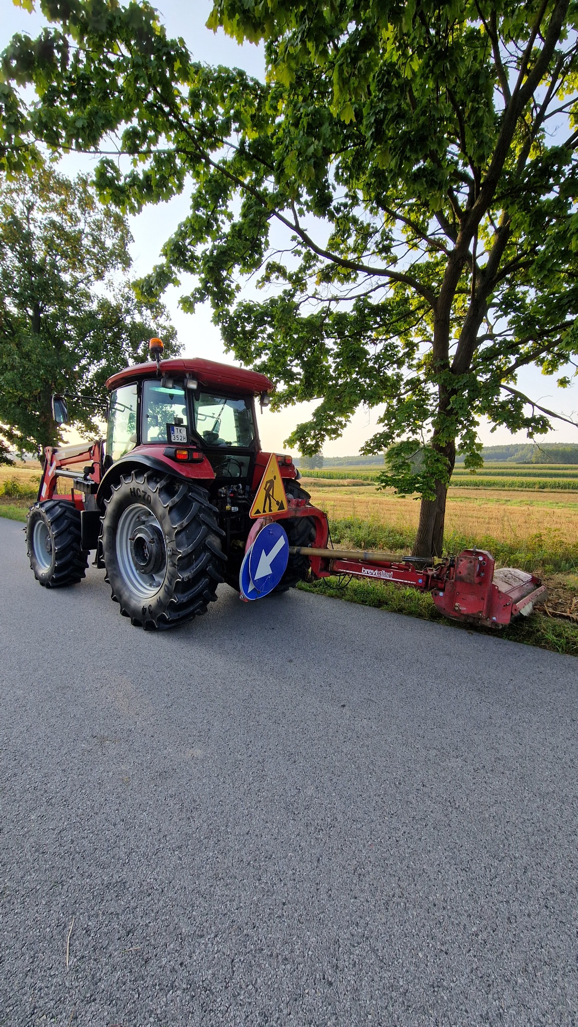 Czerwony traktor z kosiarką bijakową i znakami drogowymi na poboczu asfaltowej drogi. W tle drzewa i pole.