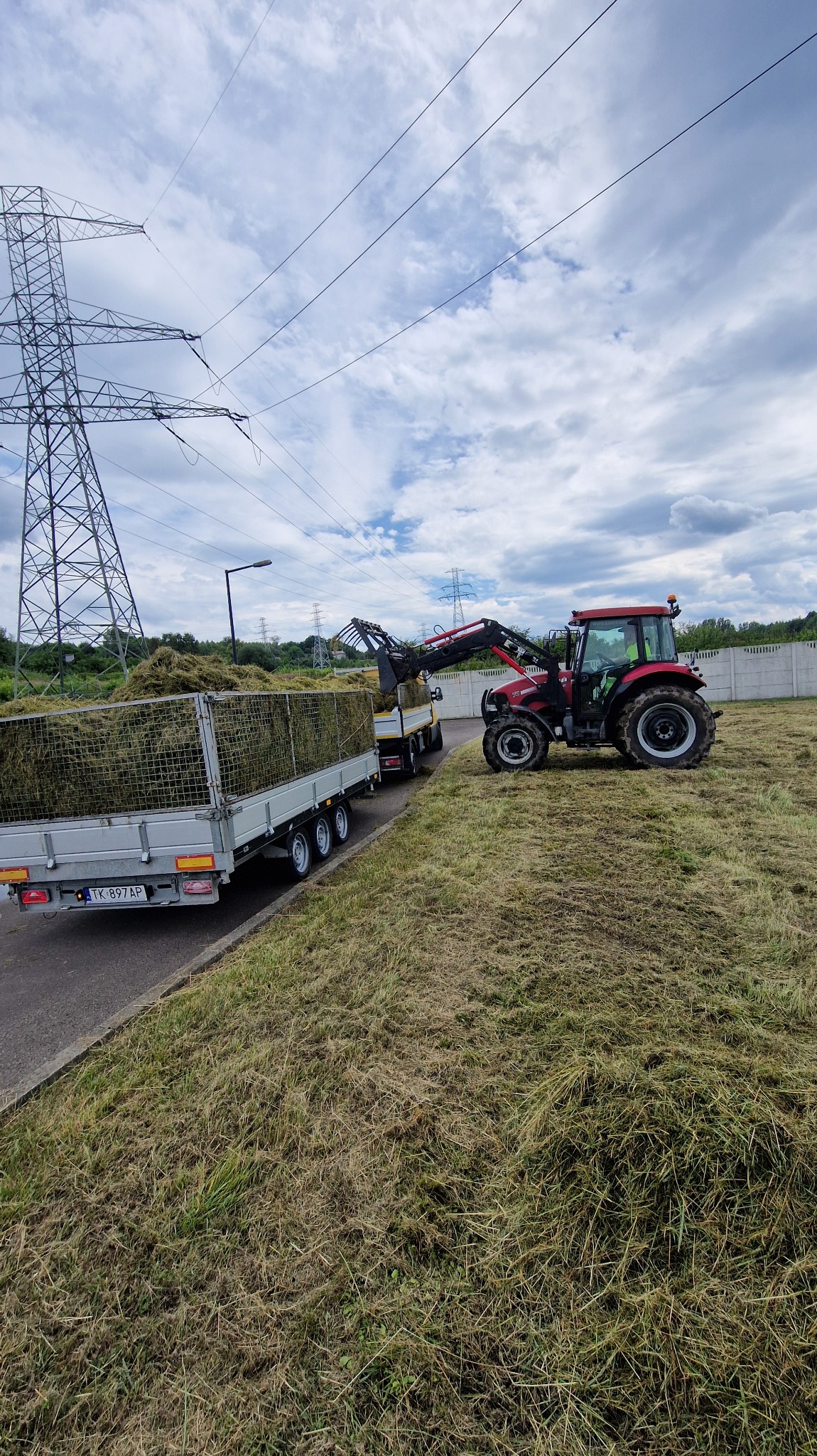 Czerwony traktor z przyczepą załadowaną skoszoną trawą na polu. W tle słupy wysokiego napięcia i fragment betonowego ogrodzenia. Praca w toku.
