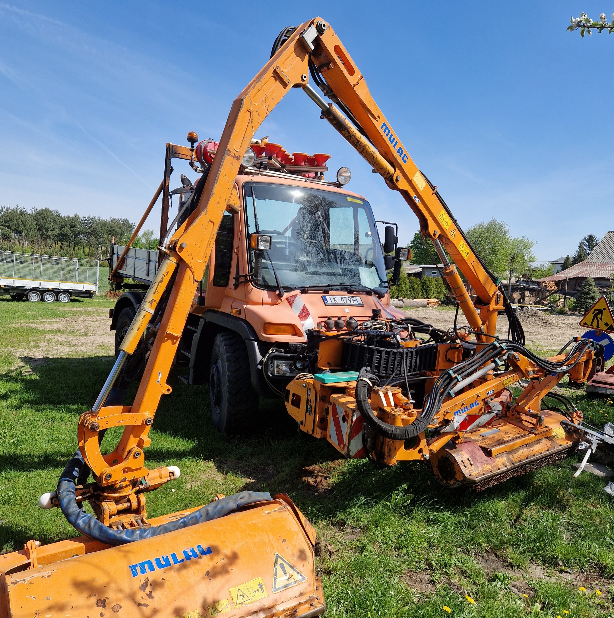 Pomarańczowy ciągnik rolniczy Mulag z wysięgnikiem i kosiarką rotacyjną na trawiastym terenie w słoneczny dzień. Widoczne detale konstrukcyjne i tablica rejestracyjna.