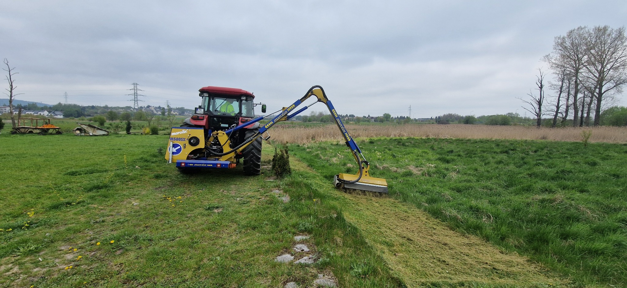 Czerwony traktor z wysięgnikiem i kosiarką rotacyjną Marolin M580SX na zielonym polu po koszeniu. Widoczne ślady koszenia i fragmenty trawy.