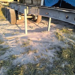 Renowacja PMS - Szarosrebrna rama naczepy z kołami, podparta na metalowych nogach, stoi na piaszczystym podłożu z trawą. W tle bele siana i namiot.