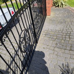 Renowacja PMS - Czarna, metalowa brama z dekoracyjnymi elementami, prowadząca do posesji z brukowanym podjazdem. Widoczny fragment muru z czerwonej cegły i roślinność.