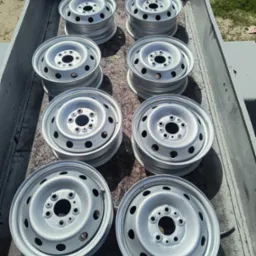 Osiem srebrnych felg samochodowych ułożonych w metalowej skrzyni, po piaskowaniu i malowaniu, widoczne z góry, z delikatnym połyskiem i cieniem.