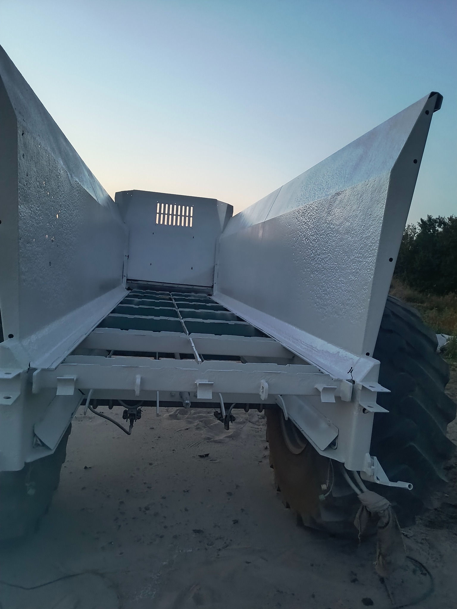 Biała, odnowiona konstrukcja metalowa maszyny rolniczej z widocznymi detalami, po piaskowaniu i malowaniu, na tle nieba o zmierzchu.