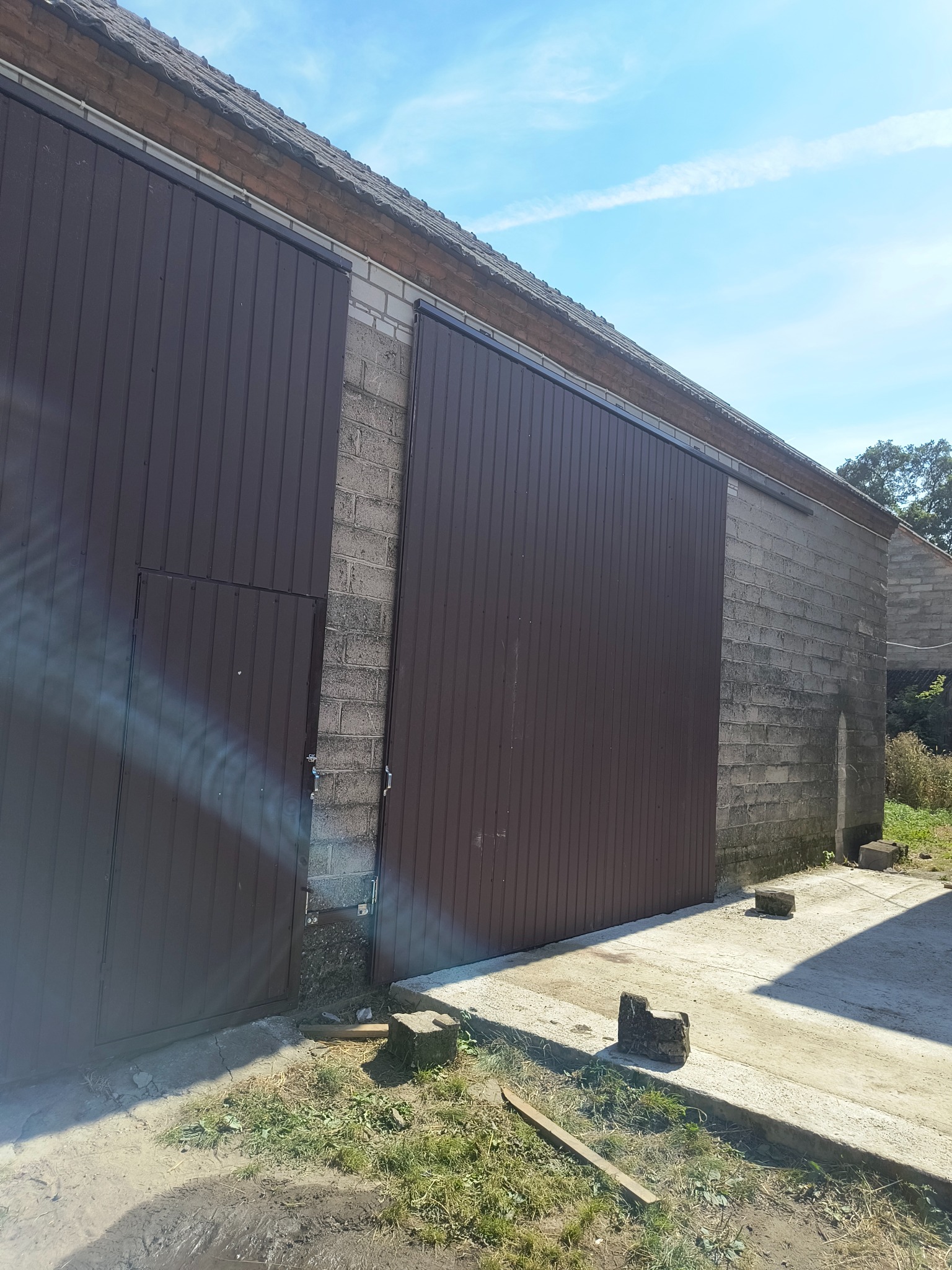 Duża, brązowa brama przesuwna z małym wejściem, zamontowana na szarym, murowanym budynku gospodarczym w słoneczny dzień. Widoczne detale konstrukcyjne i elementy otoczenia.