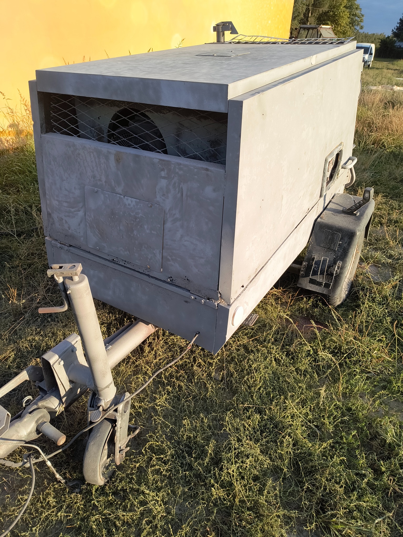 Mobilna piaskarka na przyczepie, z widoczną kratą wentylacyjną i kołami, zaparkowana na trawie. Urządzenie po renowacji, gotowe do użycia.