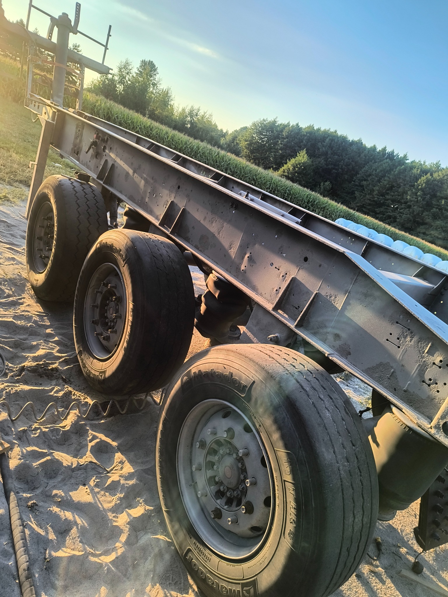 Rama naczepy z kołami na piaszczystym podłożu, widoczna z boku pod kątem, oświetlona promieniami zachodzącego słońca. Metalowa konstrukcja z widocznymi śladami użytkowania.