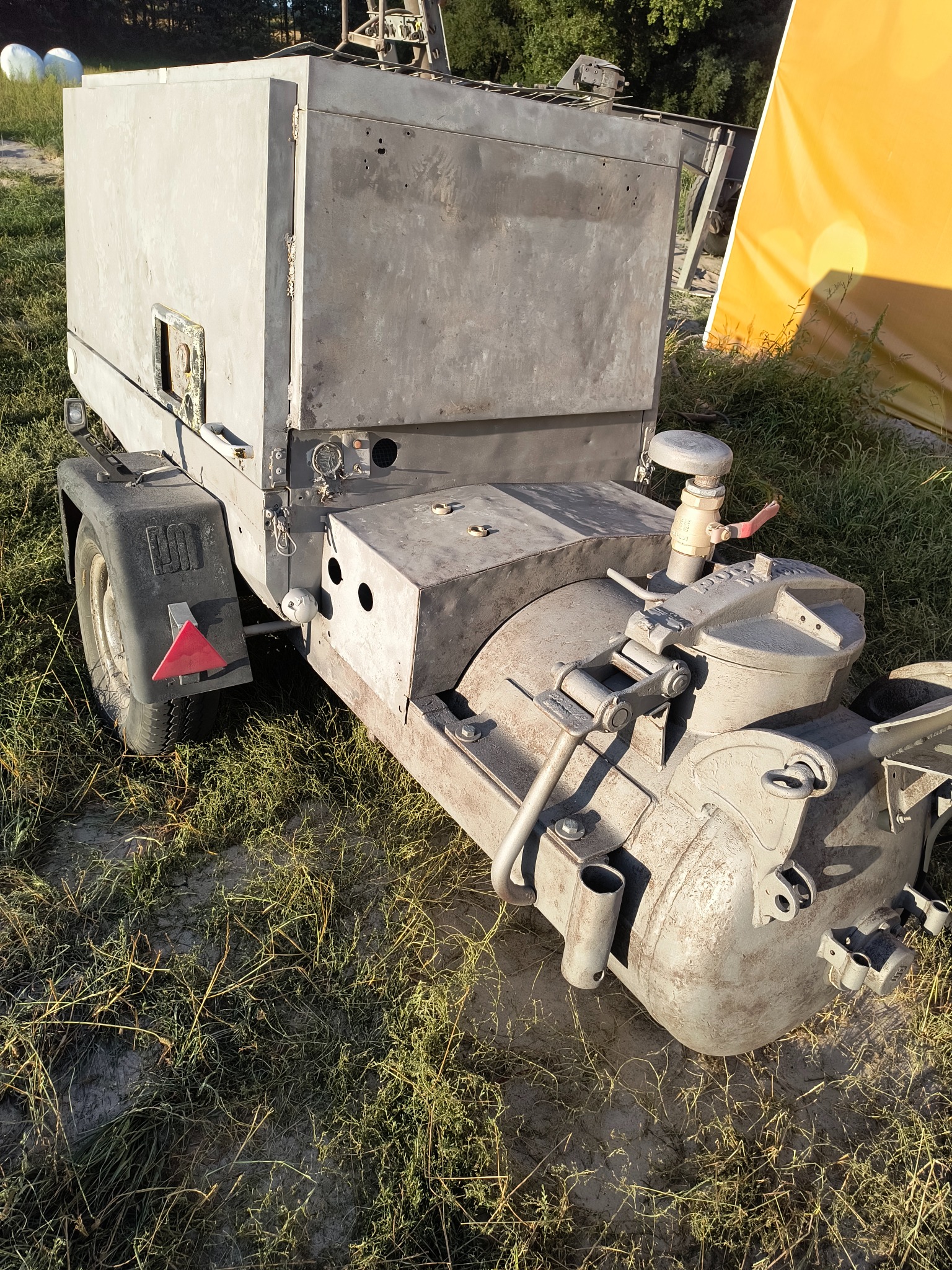 Mobilna piaskarka na przyczepce, z widocznymi elementami roboczymi i zbiornikiem, stojąca na trawiastym terenie w słoneczny dzień.