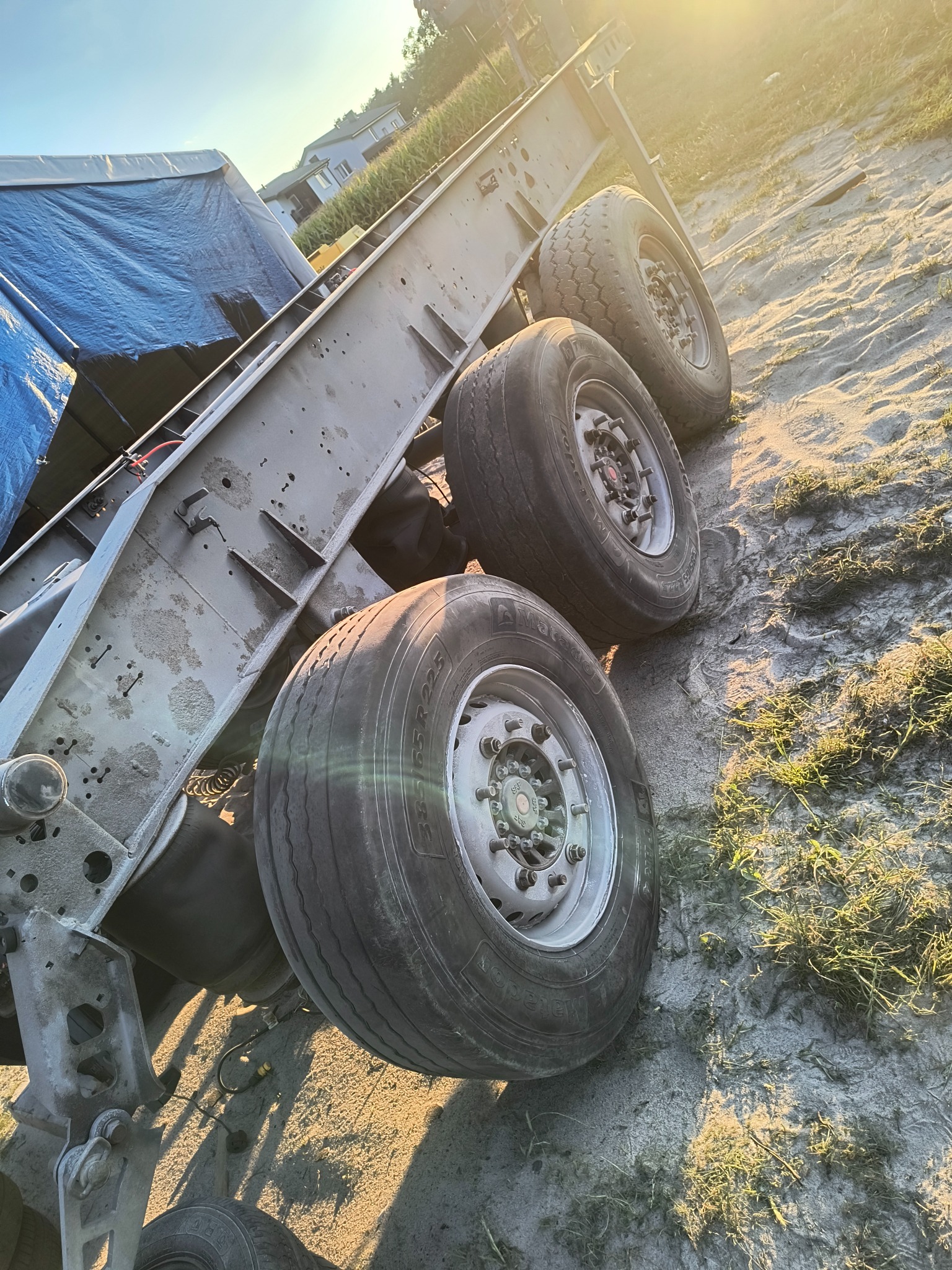 Podwozie przyczepy z trzema osiami i kołami na piaszczystym podłożu, widoczne ślady użytkowania i zabrudzenia. Oświetlenie słoneczne, kąt ujęcia z dołu.