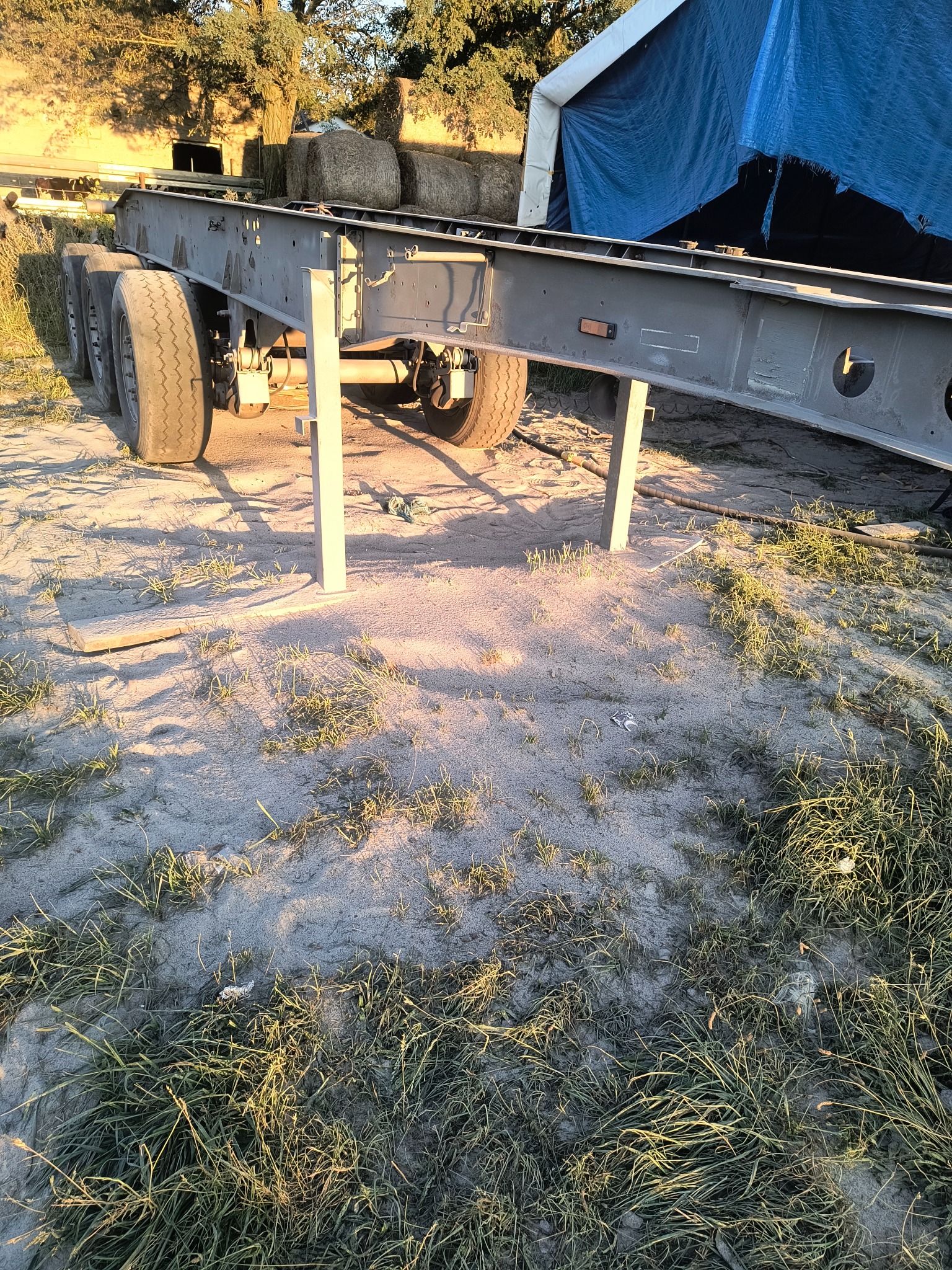 Szarosrebrna rama naczepy z kołami, podparta na metalowych nogach, stoi na piaszczystym podłożu z trawą. W tle bele siana i namiot.