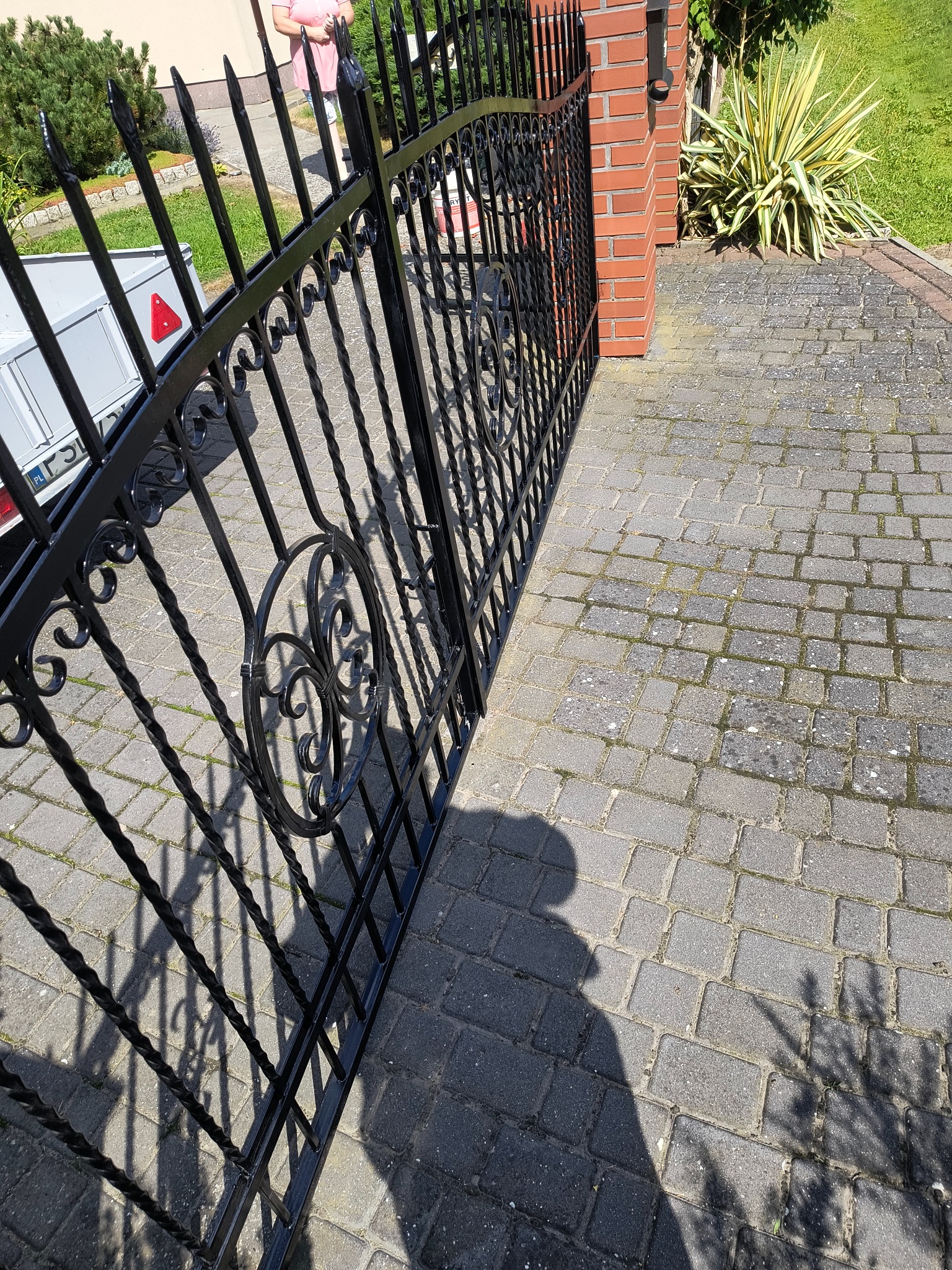Czarna, metalowa brama z dekoracyjnymi elementami, prowadząca do posesji z brukowanym podjazdem. Widoczny fragment muru z czerwonej cegły i roślinność.