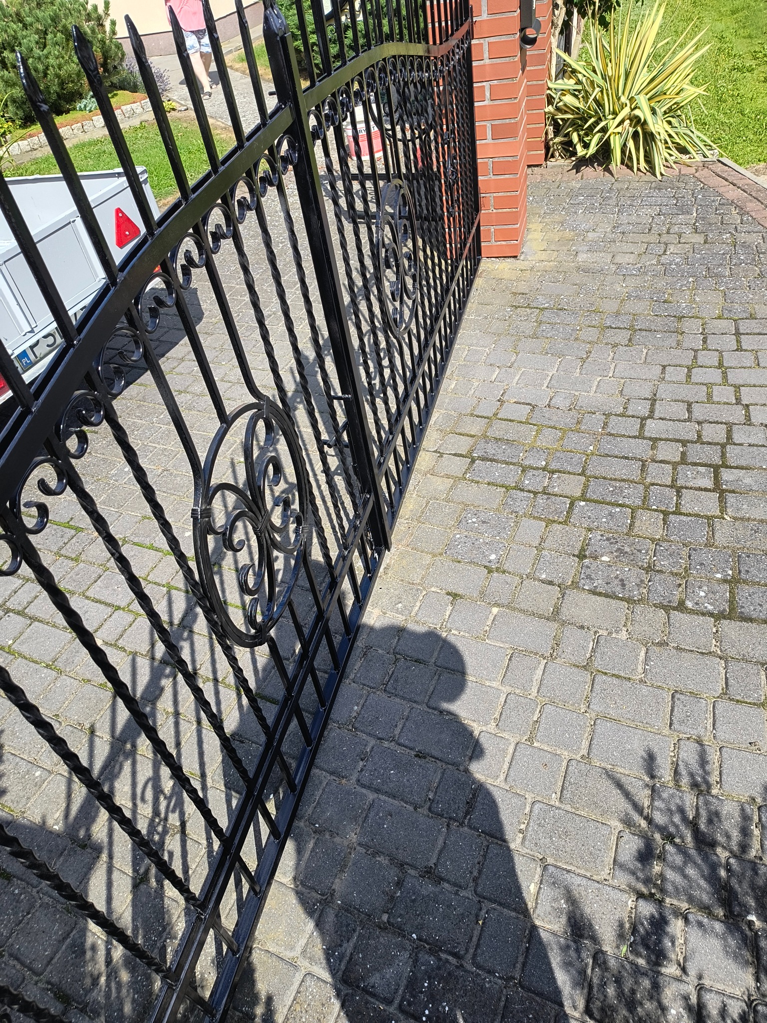 Otwarta, czarna, metalowa brama z dekoracyjnymi elementami, prowadząca na wybrukowany podjazd. Widoczny fragment ceglanego słupka i roślinność w tle.
