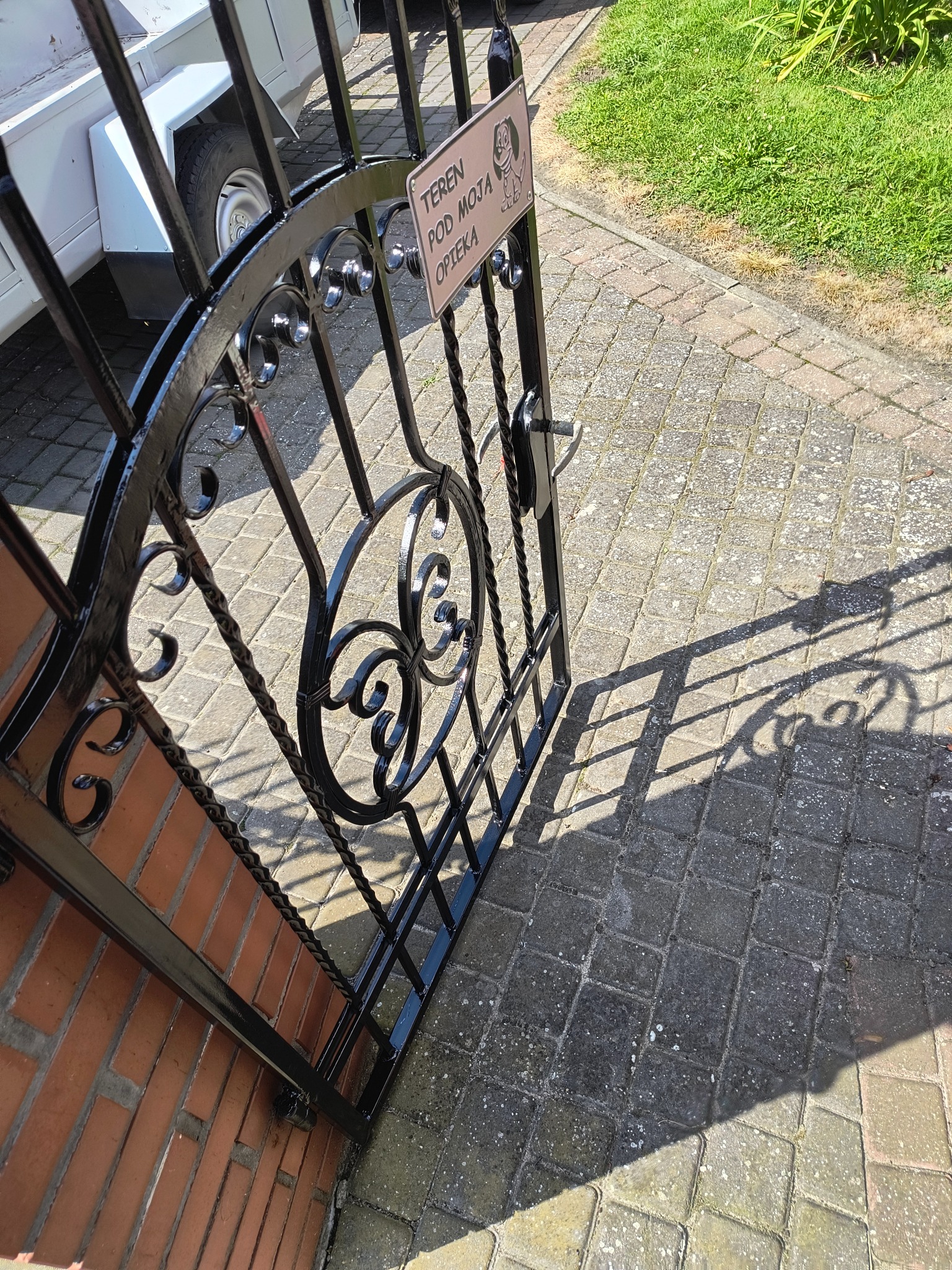 Otwarta, czarna, metalowa furtka z ozdobnymi elementami i tabliczką informacyjną, osadzona w murze z cegły, z widocznym cieniem na brukowanym podłożu.