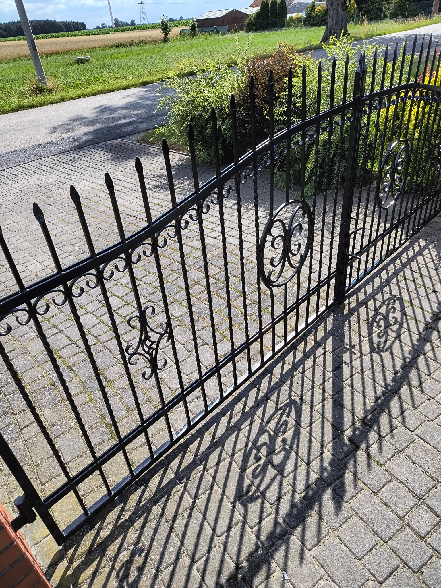 Kute, czarne ogrodzenie metalowe z dekoracyjnymi elementami i cieniem na brukowanej nawierzchni. Widok na zielony ogród i drogę w tle.