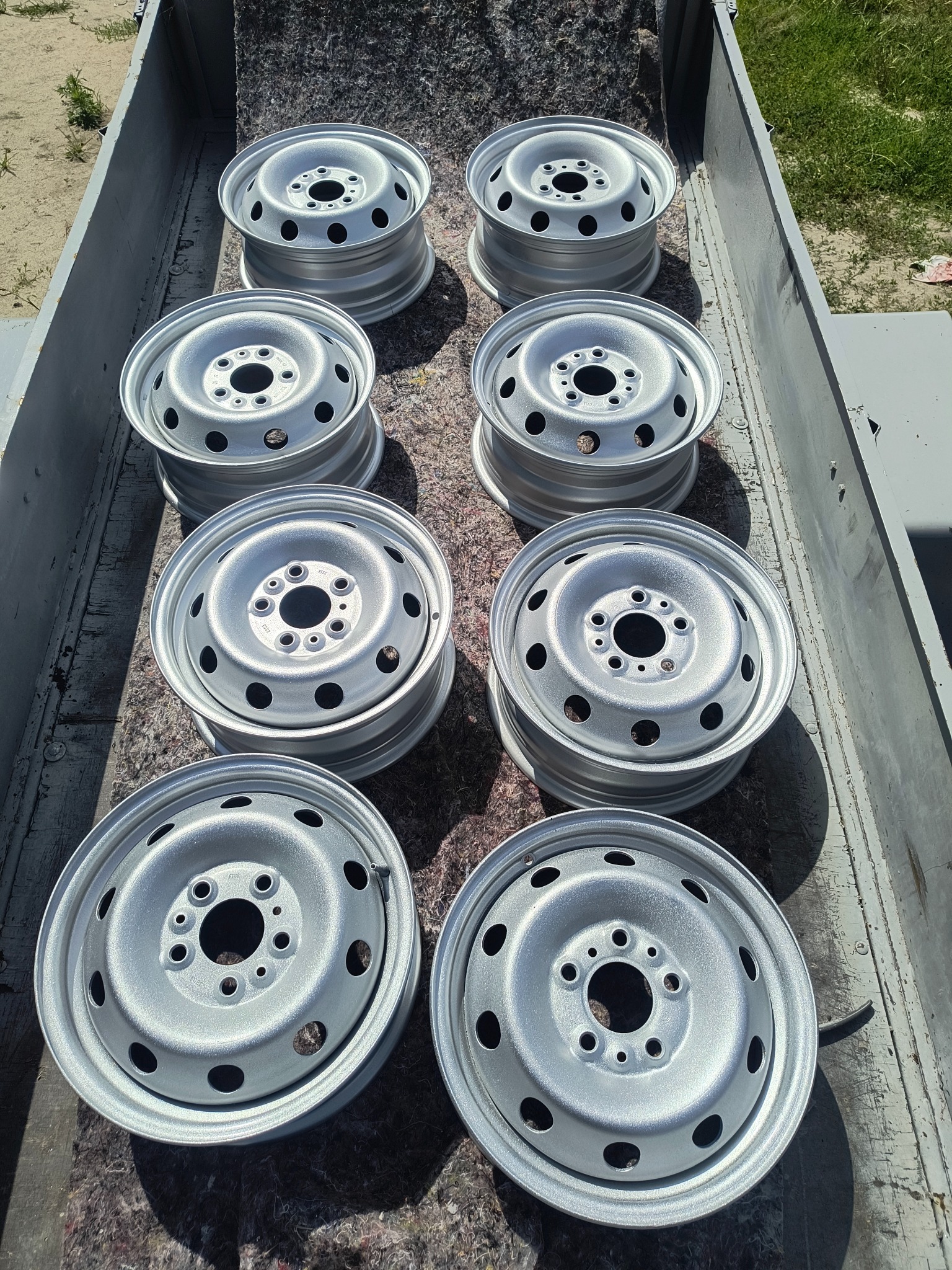 Osiem srebrnych felg samochodowych ułożonych w metalowej skrzyni, po piaskowaniu i malowaniu, widoczne z góry, z delikatnym połyskiem i cieniem.