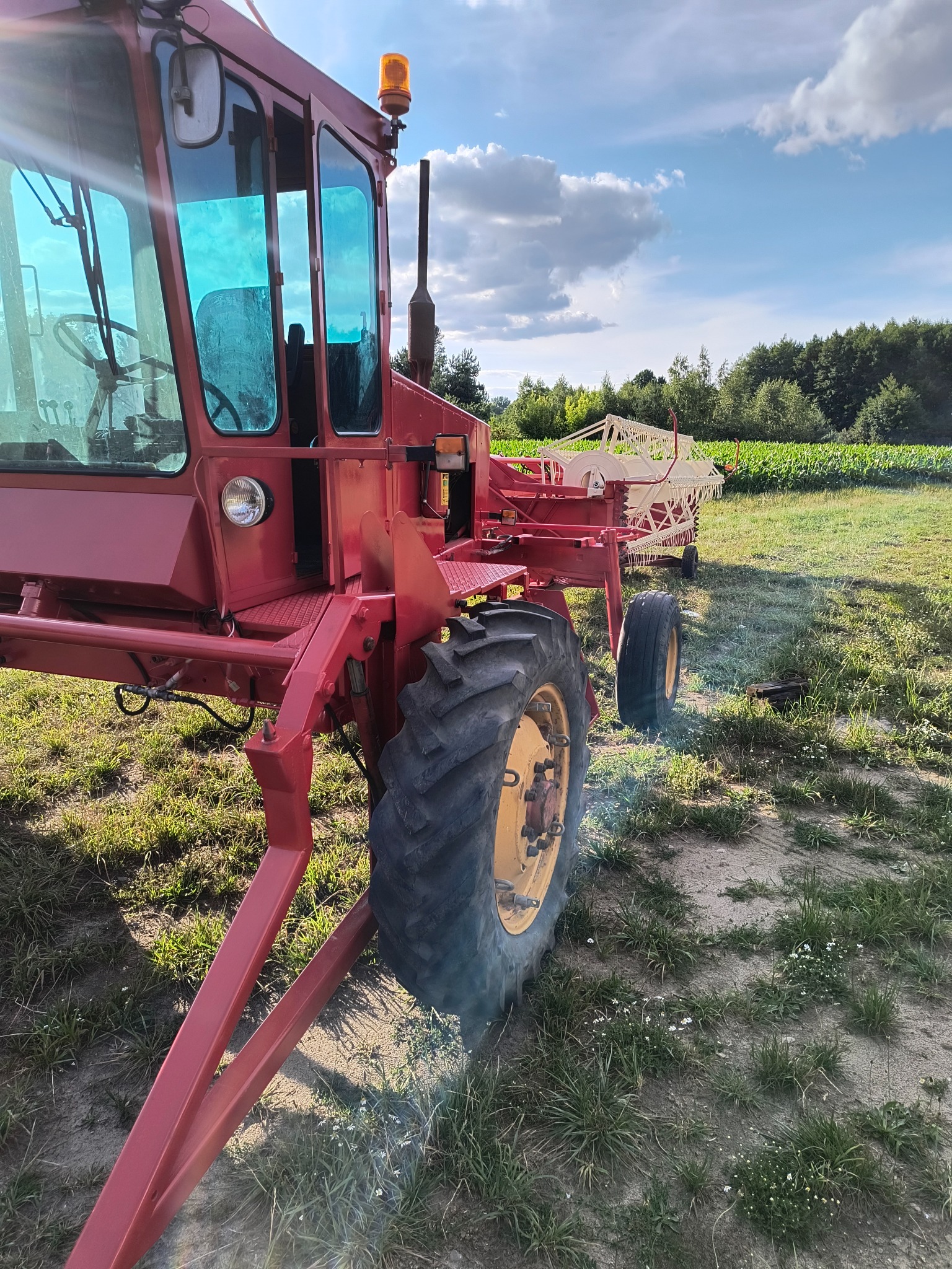 Czerwony kombajn rolniczy z żółtymi felgami na polu z widocznym niebem i drzewami w tle. Detal kabiny i koła. Specyficzny model maszyny rolniczej.