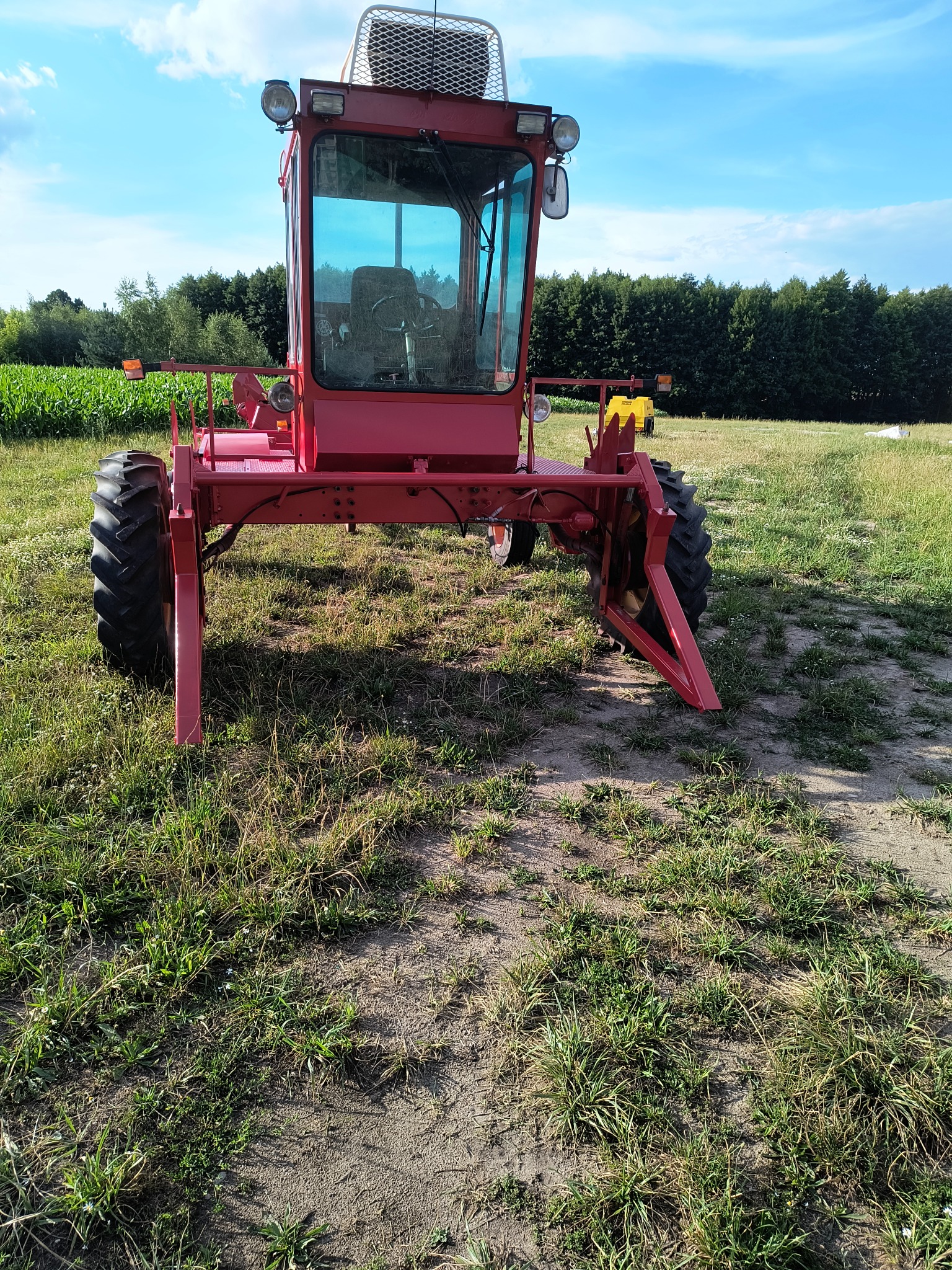 Czerwony, specjalistyczny pojazd rolniczy z kabiną, na polnej drodze, z widocznymi elementami roboczymi i oponami z bieżnikiem, na tle zieleni i błękitnego nieba.