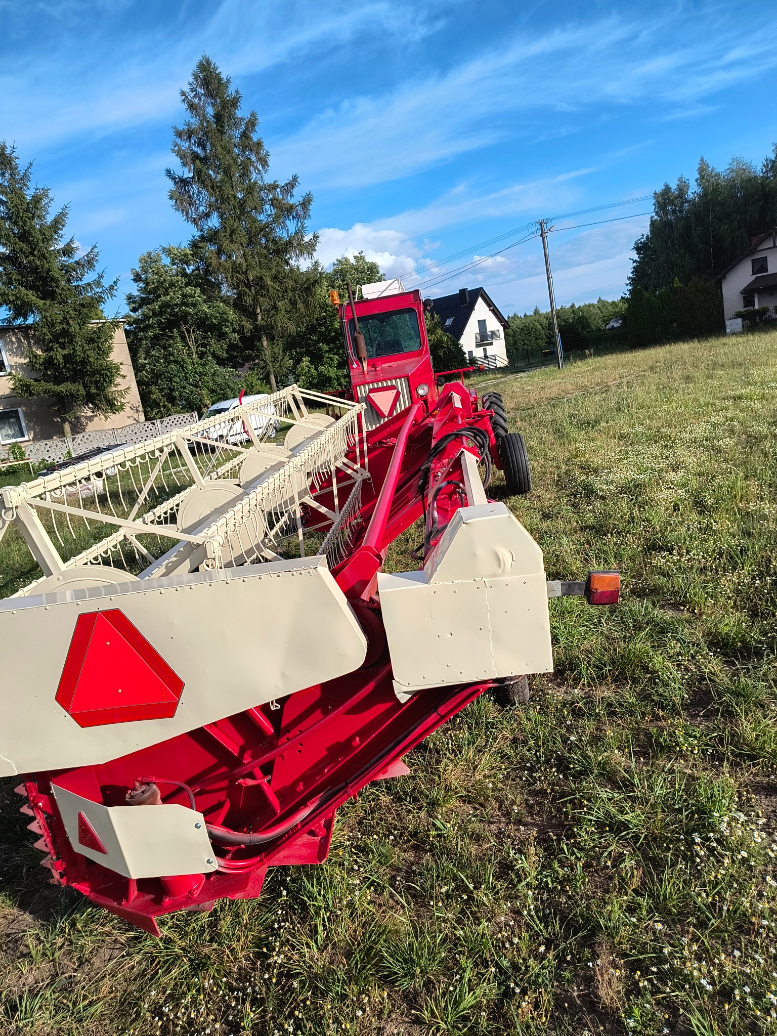 Czerwono-biały heder kombajnu na łące, z widocznymi detalami konstrukcyjnymi i elementami tnącymi. W tle domy i błękitne niebo.
