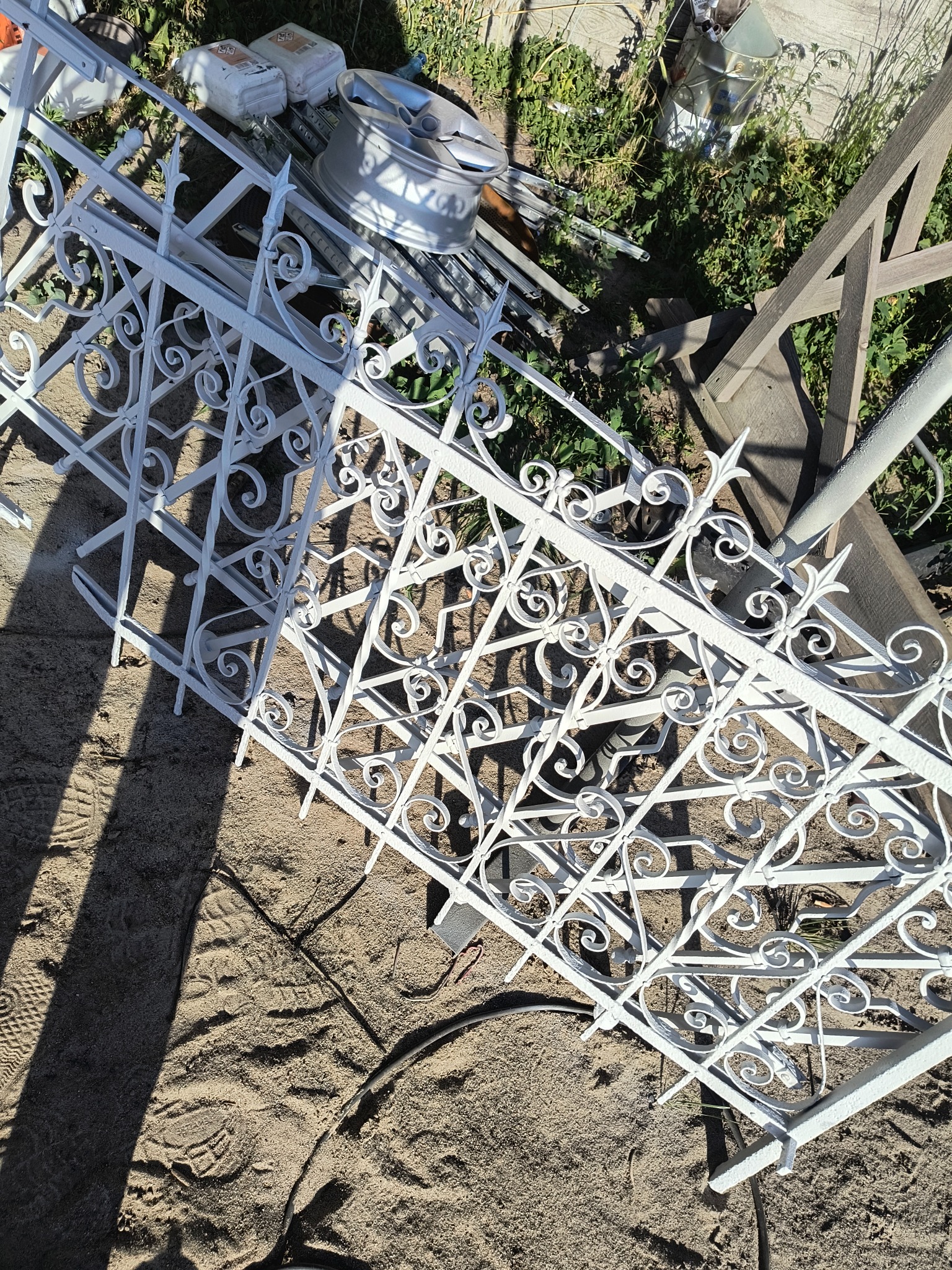 Biały, ozdobny fragment ogrodzenia metalowego z dekoracyjnymi elementami leżący na piasku, z widocznymi śladami butów i elementami otoczenia w tle.