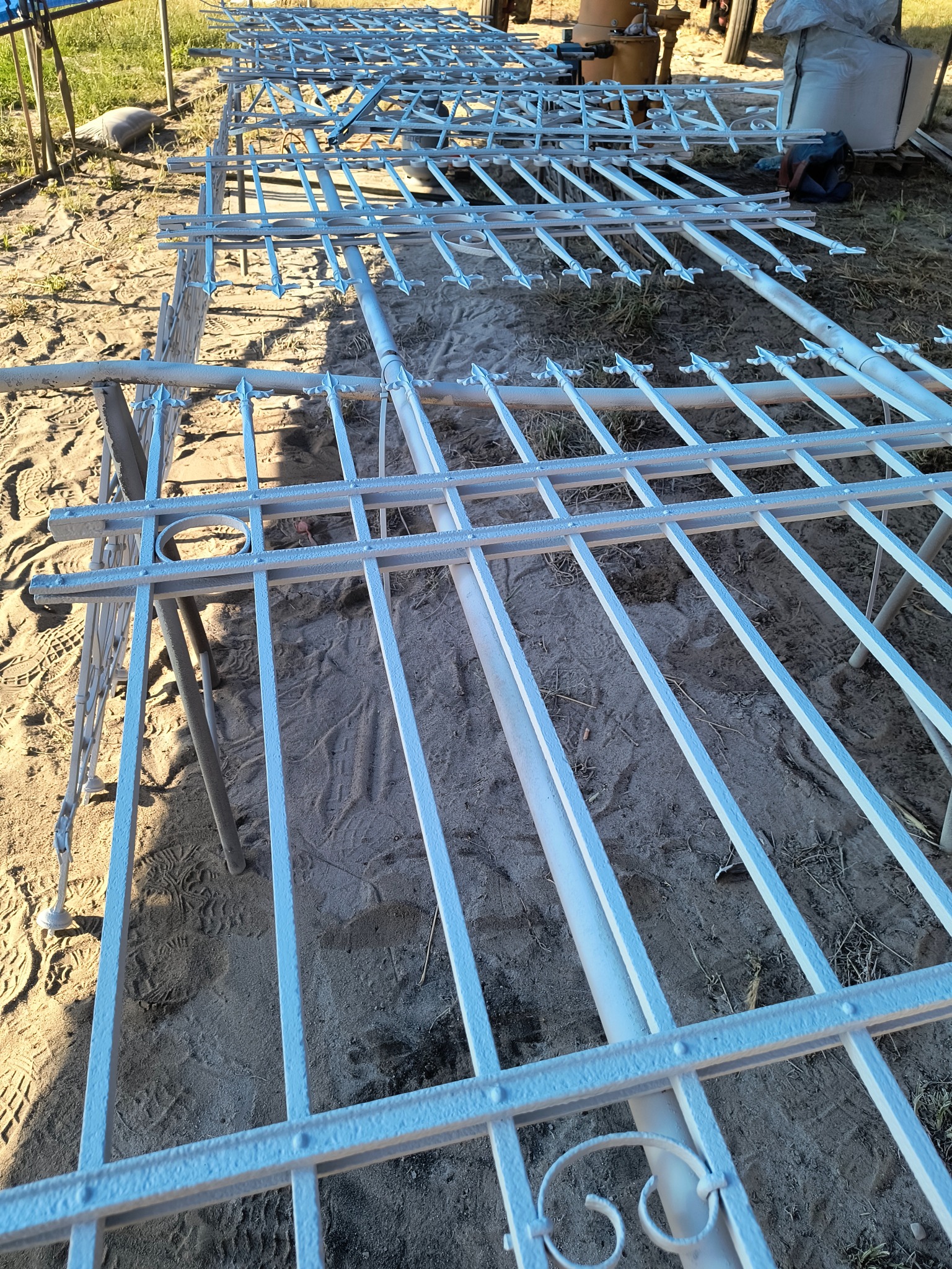 Białe, metalowe elementy ogrodzenia z dekoracyjnymi grotami, rozłożone na piasku po malowaniu, gotowe do montażu. Widoczne ślady stóp na piasku.