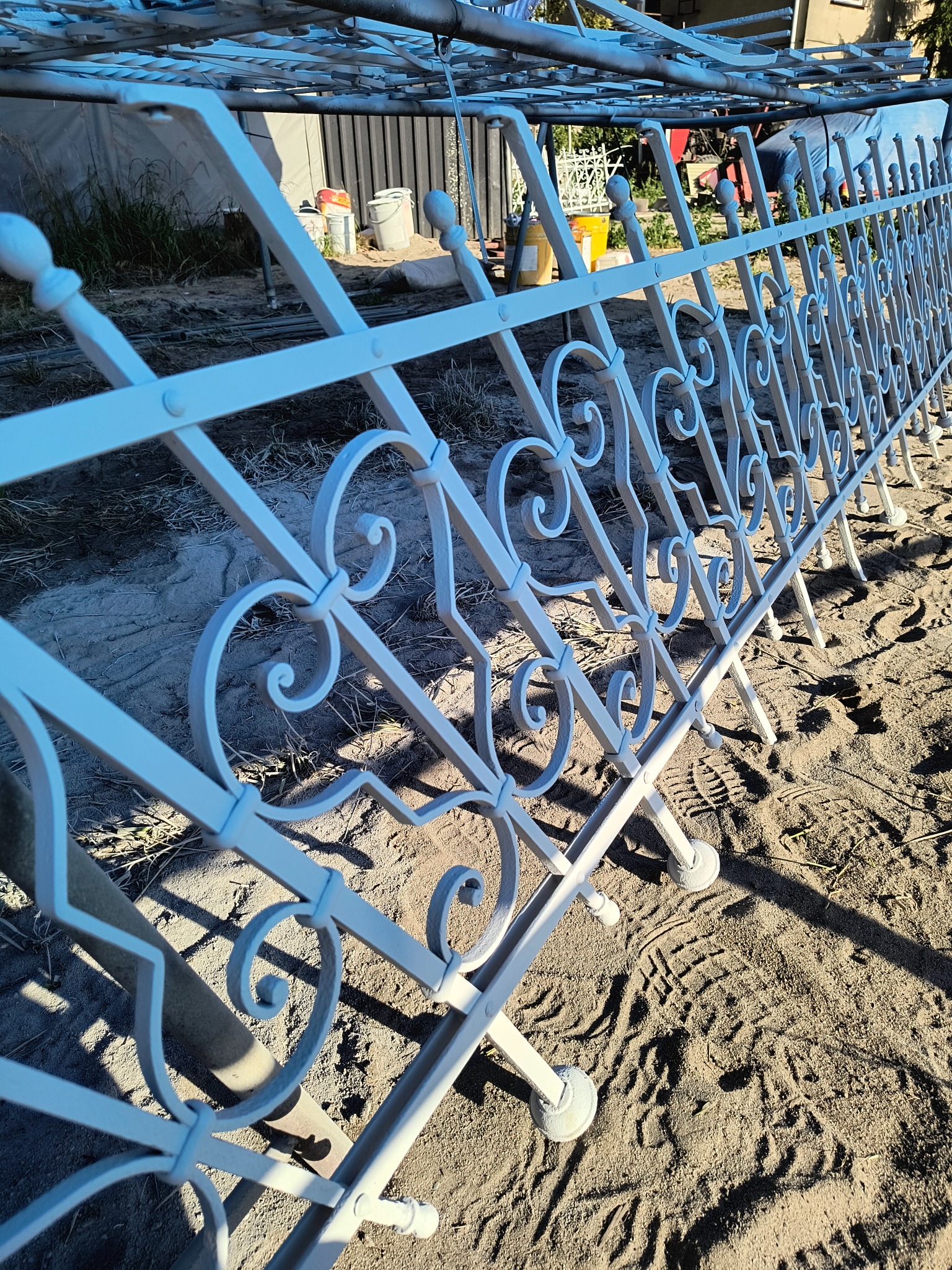 Białe, dekoracyjne ogrodzenie metalowe z ozdobnymi elementami w kształcie serc, świeżo po malowaniu, ustawione na piasku. Widoczne ślady butów.