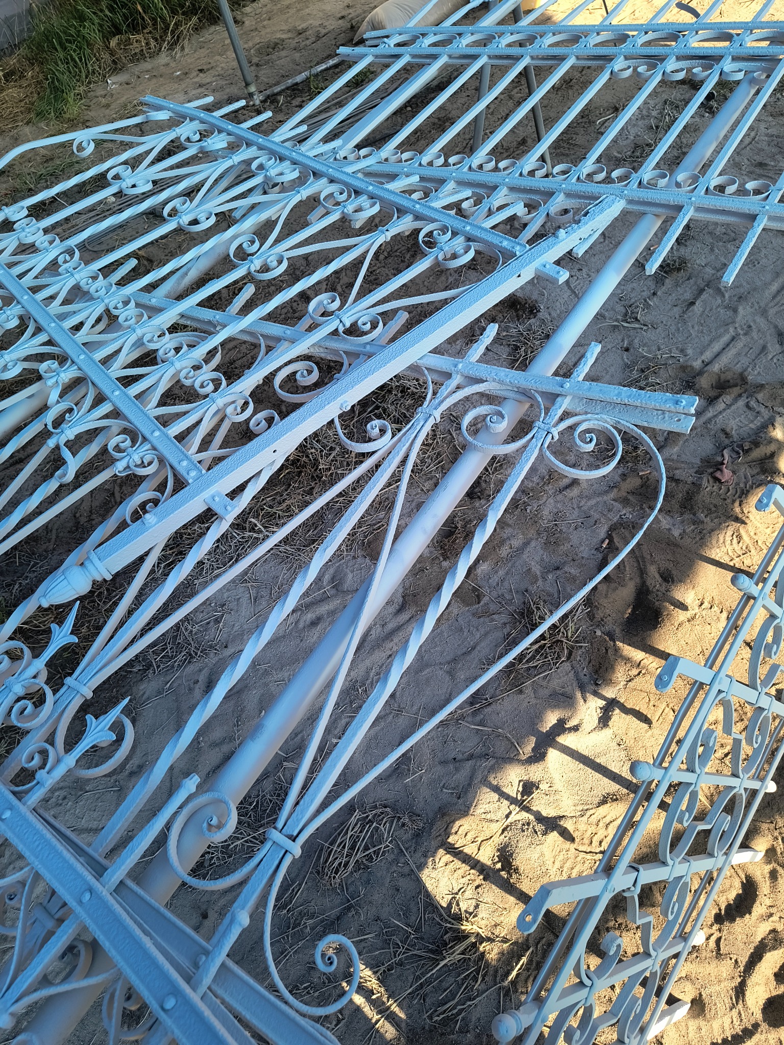 Białe, metalowe elementy dekoracyjne, prawdopodobnie balustrady lub ogrodzenia, leżące na piasku. Widoczne detale i wzory.