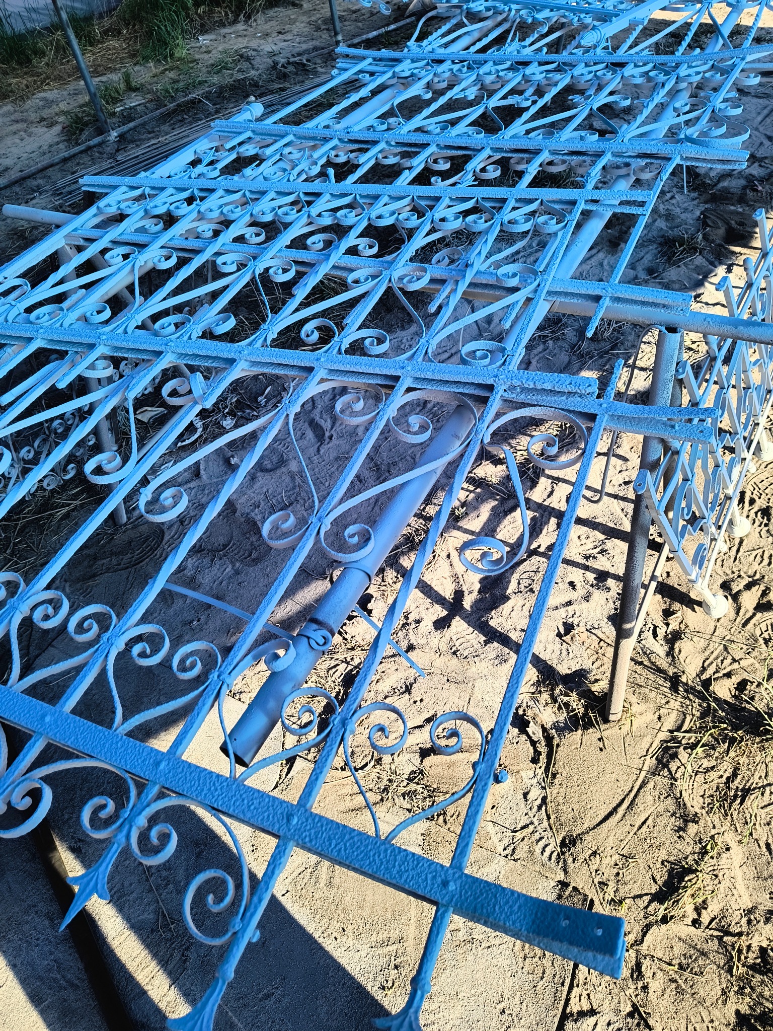 Błękitne, dekoracyjne elementy metalowe, prawdopodobnie fragmenty ogrodzenia, leżące na ziemi. Widoczna faktura po malowaniu, z bliska, w słoneczny dzień.