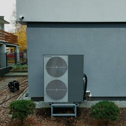PanHydraulik - Szara pompa ciepła Technix zamontowana na metalowym stelażu, przy ścianie budynku z kamerą monitoringu. Obok małe drzewka iglaste.
