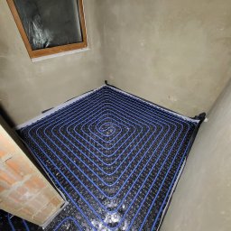 PanHydraulik - Niebieskie rury ogrzewania podłogowego ułożone spiralnie na czarnej macie w surowym pomieszczeniu z oknem i fragmentem cegły. Instalacja przed wylaniem posadzki.