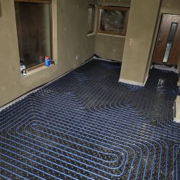 PanHydraulik - Niebieskie rury ogrzewania podłogowego ułożone spiralnie na czarnej macie w surowym wnętrzu z oknami i drzwiami, gotowe do wylania posadzki.